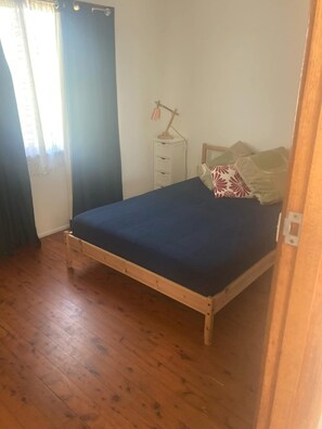 Uma cama de solteiro com estrutura de madeira e colchão azul escuro em quarto com piso de madeira.