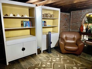Una habitación con una butaca de cuero marrón, estanterías blancas con libros y adornos, un cesto de basura y una alfombra estampada.