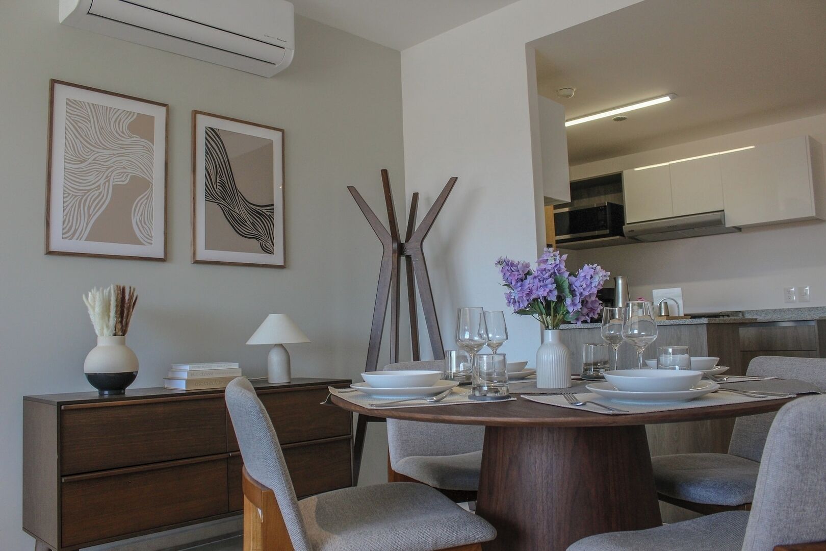 Un coin repas moderne avec une table ronde dressée pour quatre personnes, entourée de chaises à revêtement gris. Deux œuvres d’art abstraites encadrées ornent le mur, un vase avec des fleurs violettes est posé sur la table, et une lampe blanche se trouve à côté de la table.