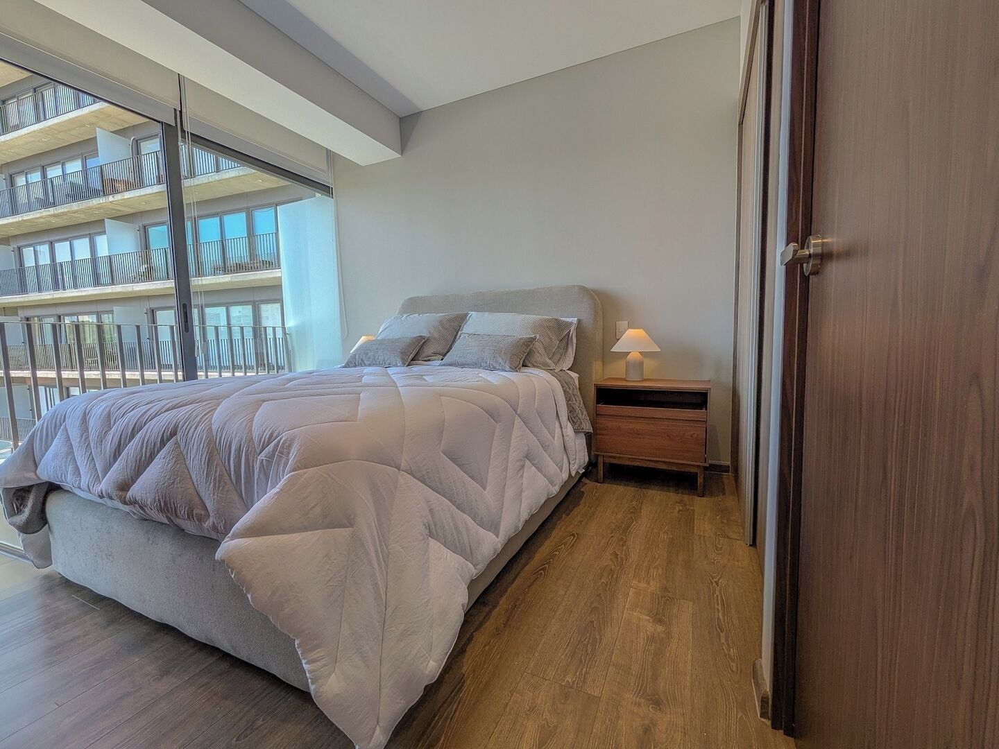 A bedroom with a bed, bedside table, and a view of a balcony.