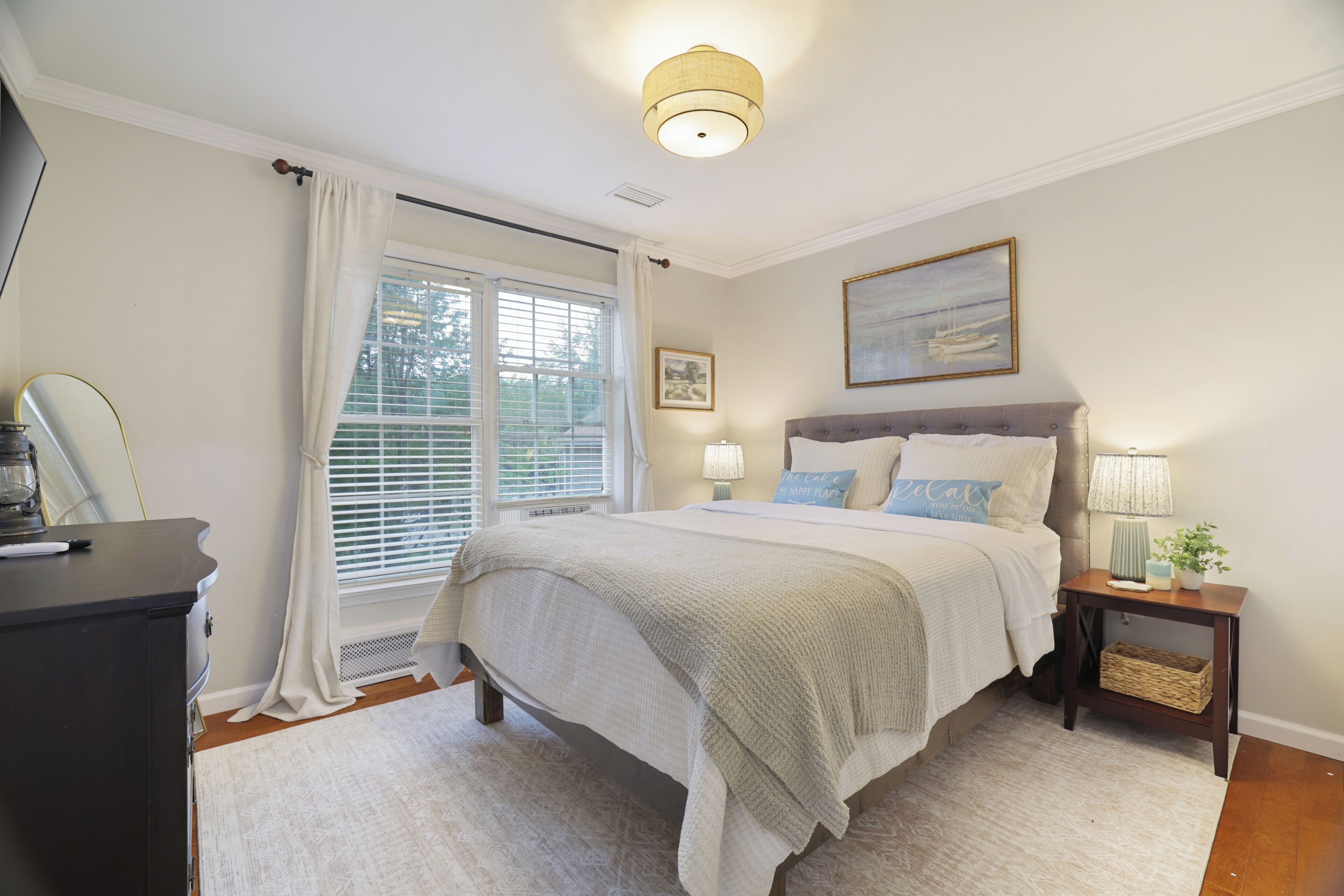 Un dormitorio con cama, mesitas de noche, lámpara, un cuadro y ventana con cortinas.