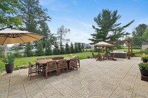 An outdoor patio with a wooden table and chairs, two umbrellas, and a hot tub. - La Belle Vie 
Hot Tub • Fire Pit • Outdoor Kitchen • Sleeps 12 (Lake Ariel)