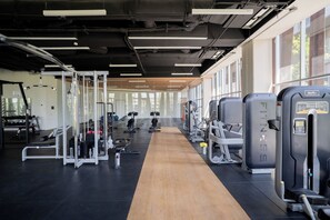 Ein Fitnessraum mit unterschiedlichen Trainingsgeräten, Holzboden und großen Fenstern.