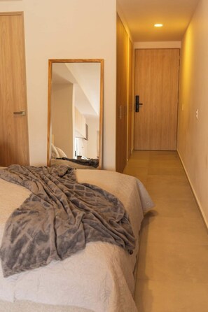 Ein Hotelzimmer mit einem Bett, einem großen Spiegel, einer Holztür und einem Flur, der zu einem weiteren Zimmer führt.