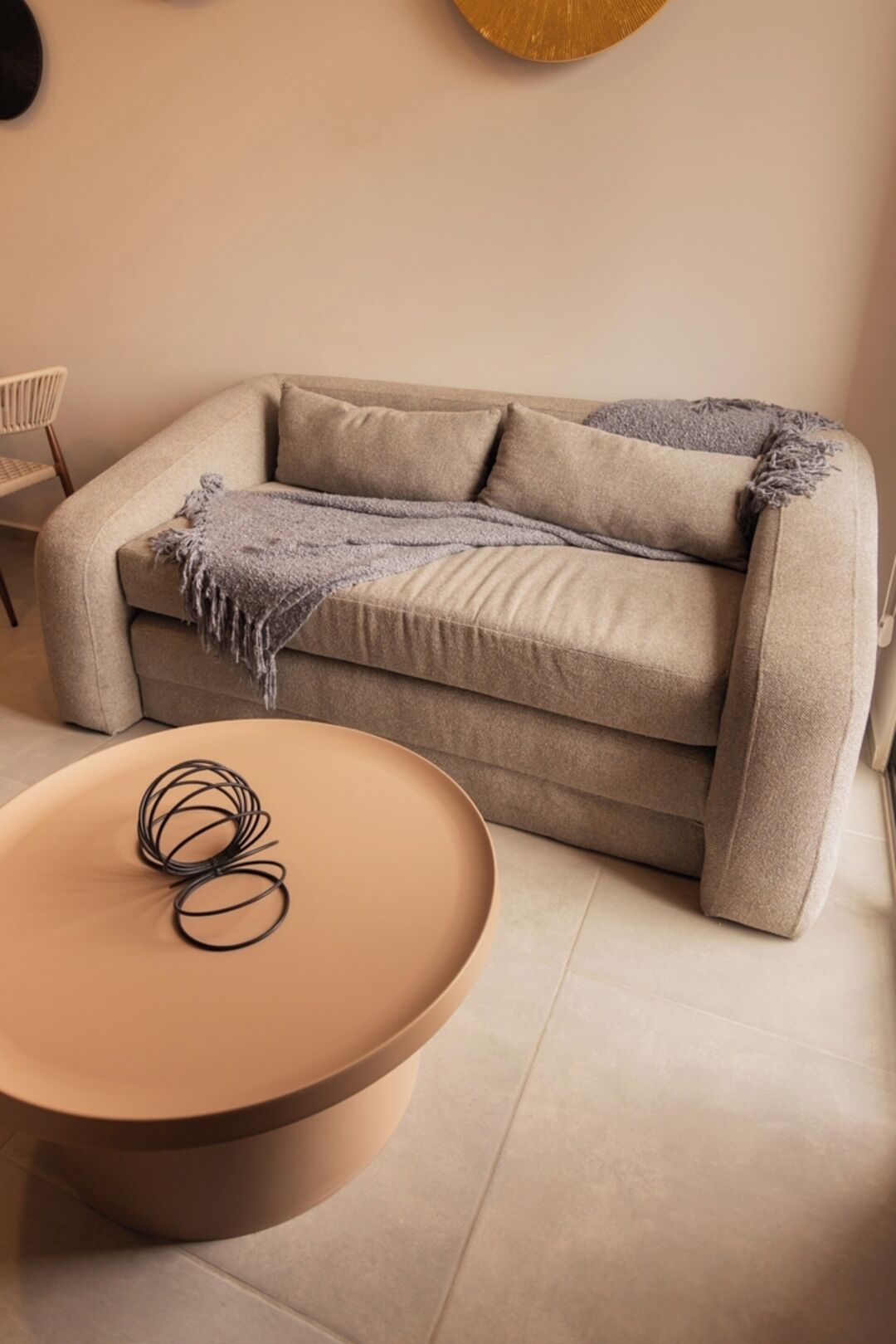 Ein beiges Sofa mit Überwurfdecke und Kissen, ein runder Couchtisch mit einem eingerollten Gegenstand und ein Sessel in einem neutral gestalteten Raum.