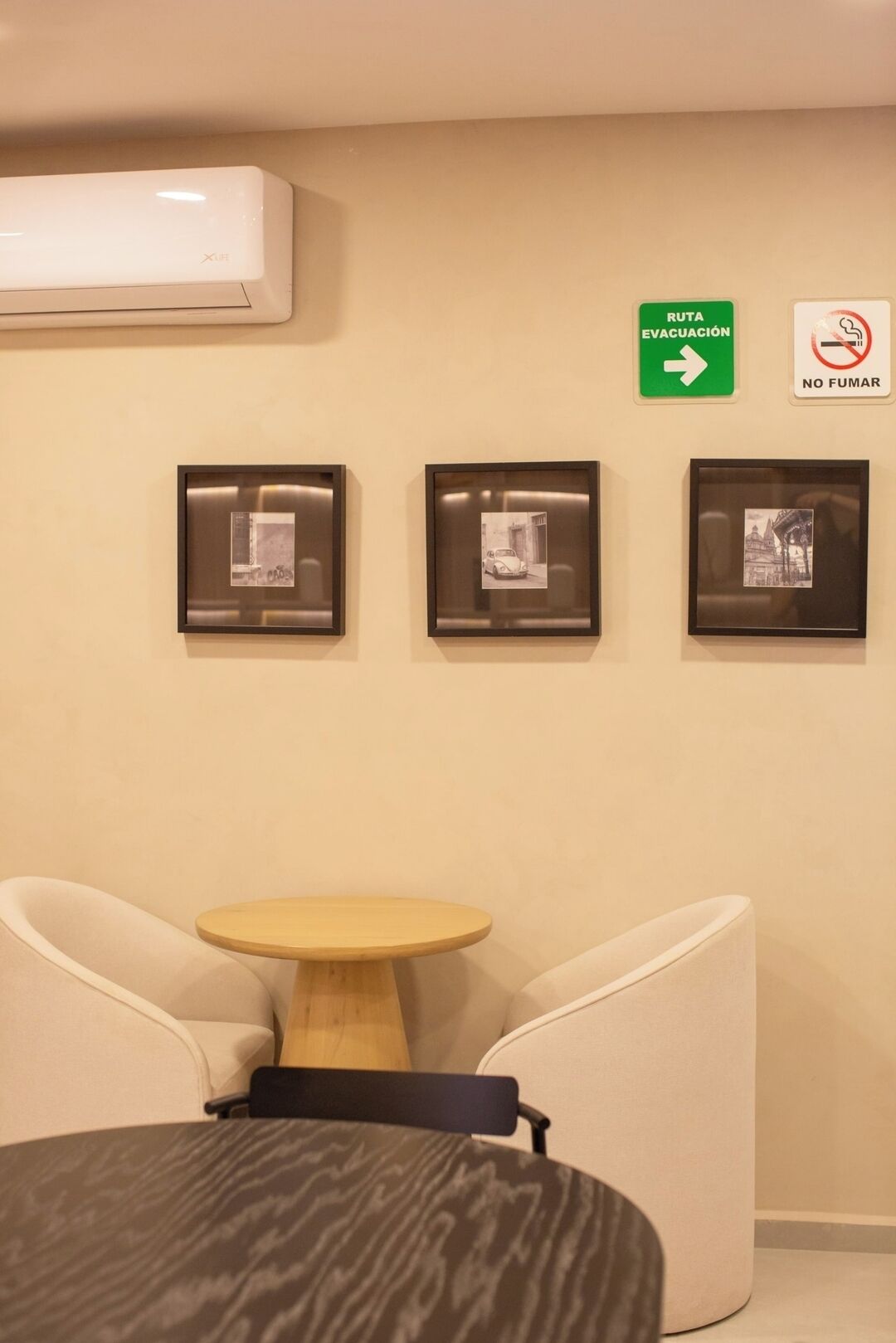 A room with a round wooden table, two chairs, and three framed pictures on the wall. There is a green evacuation route sign and a no-smoking sign.