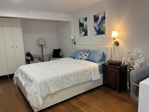 Un dormitorio con cama, mesita de noche, lámpara, sillón y una obra de arte en la pared.