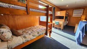 Ein Schlafraum mit Etagenbetten, einem Schreibtisch und Stuhl, einem kleinen Tisch mit blauer Tischdecke und einem Fenster mit Vorhängen.