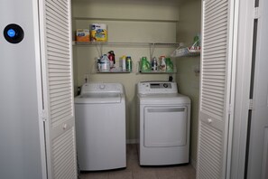 A laundry room with two white front-loading washing machines, shelves with laundry detergent, and a wall-mounted recycling bin. - Prime Location • Gym • Dual-Monitor Workspace (Tampa)