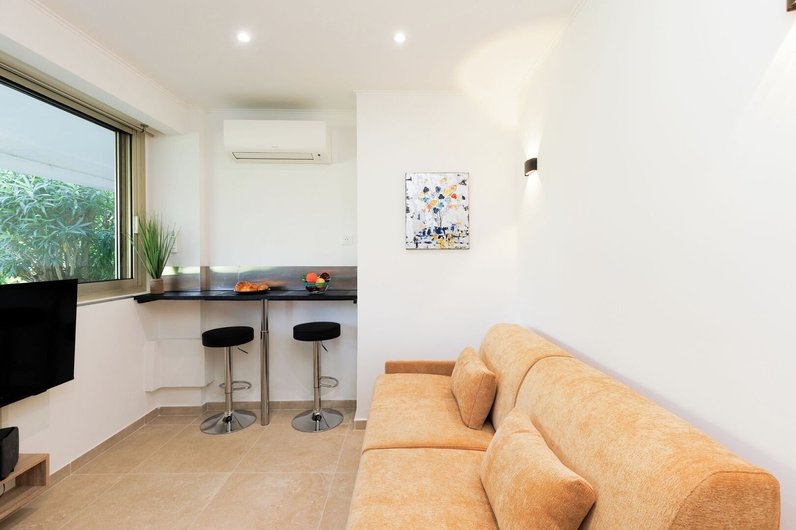 A modern living space with a sofa, a kitchen area with a counter and stools, and a window with a view of greenery.