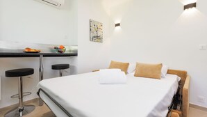 A hotel room with a bed, two bar stools, a small table with fruit, a painting, and a wall-mounted light fixture. - Upstays Cosy Studio 15m² with pool and quiet (Cannes)