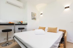 A hotel room with a bed, two bar stools, a small table with fruit, a painting, and a wall-mounted light fixture. - Upstays Cosy Studio 15m² with pool and quiet (Cannes)