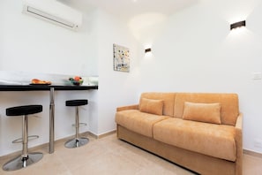 Ein modernes Wohnzimmer mit einem beigen Sofa, zwei schwarzen Barhocker, einer Küchenzeile mit Obstschale und einer wandmontierten Leuchte.