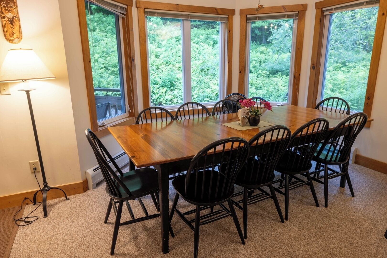 Un coin repas avec une table en bois et des chaises noires, de grandes fenêtres donnant sur un paysage verdoyant et un lampadaire.