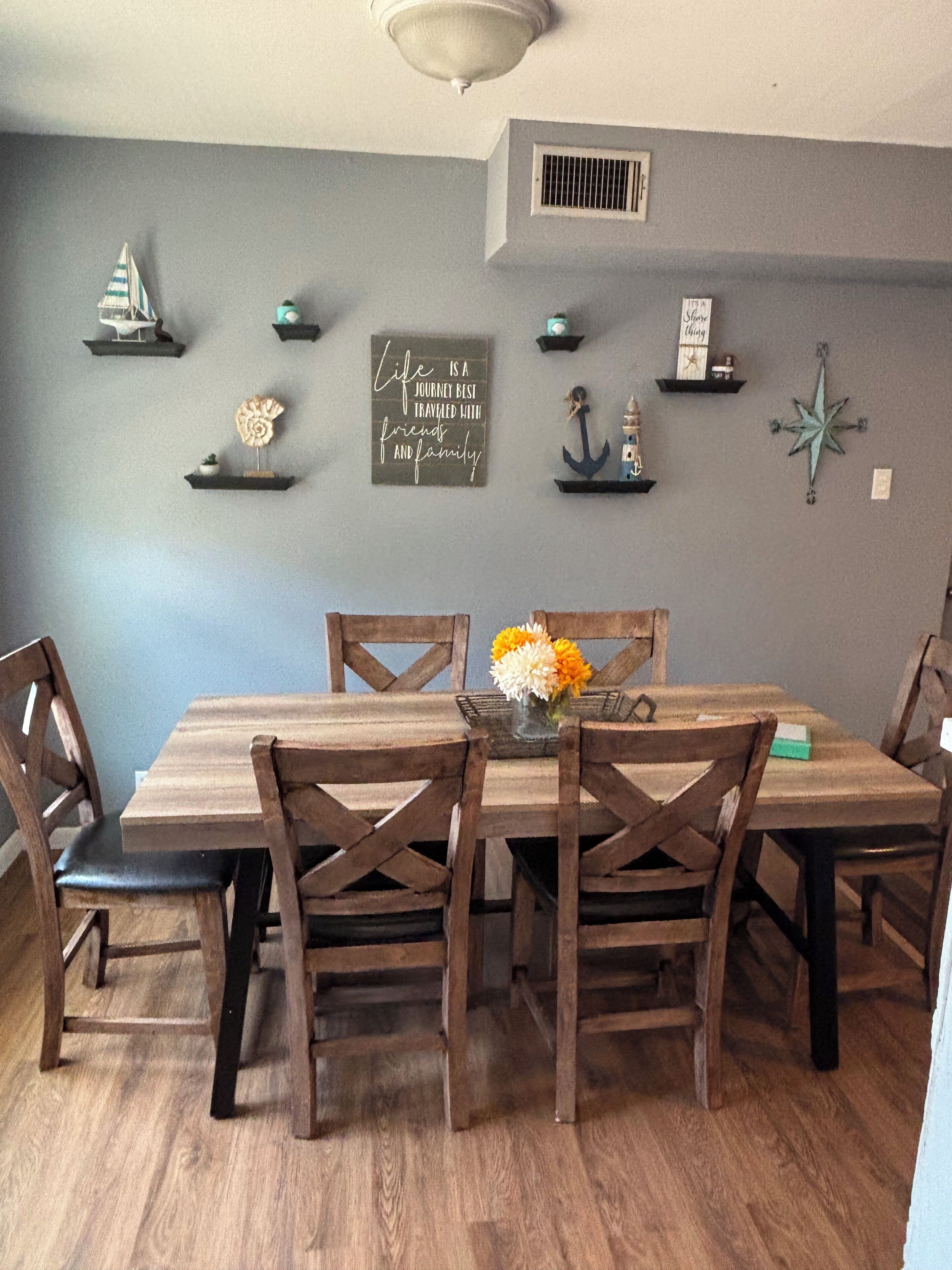 A dining area with a wooden table and chairs, a vase with flowers, and wall decorations including a compass and a sign with a quote.