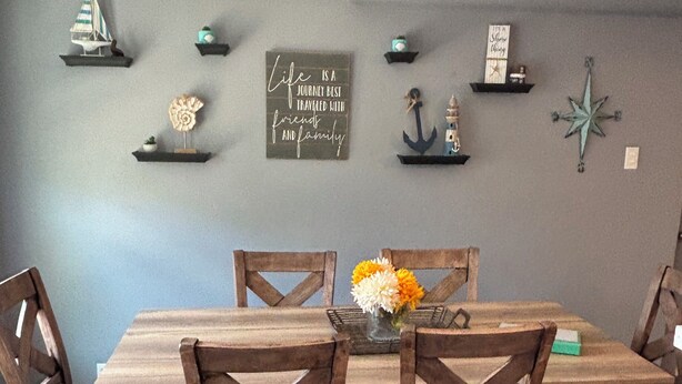A dining area with a wooden table and chairs, a vase with flowers, and wall decorations including a compass and a sign with a quote. -    Cozy Three bedroom, Family-Friendly Home with Pool, Game Room & Fast Wi-Fi (Corpus Christi)