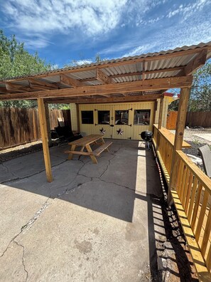 Eine überdachte Terrasse im Freien mit einem Holztisch, Bänken, einem Grill und einem Holzzaun.