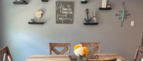 A dining area with a wooden table and chairs, a vase with flowers, and wall decorations including a compass and a sign with a quote.