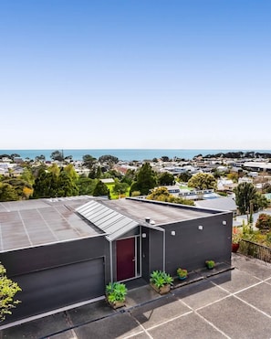A modern building with a flat roof and a red door, surrounded by greenery and a clear view of the ocean. - Views on Devon West - New Plymouth (New Plymouth)