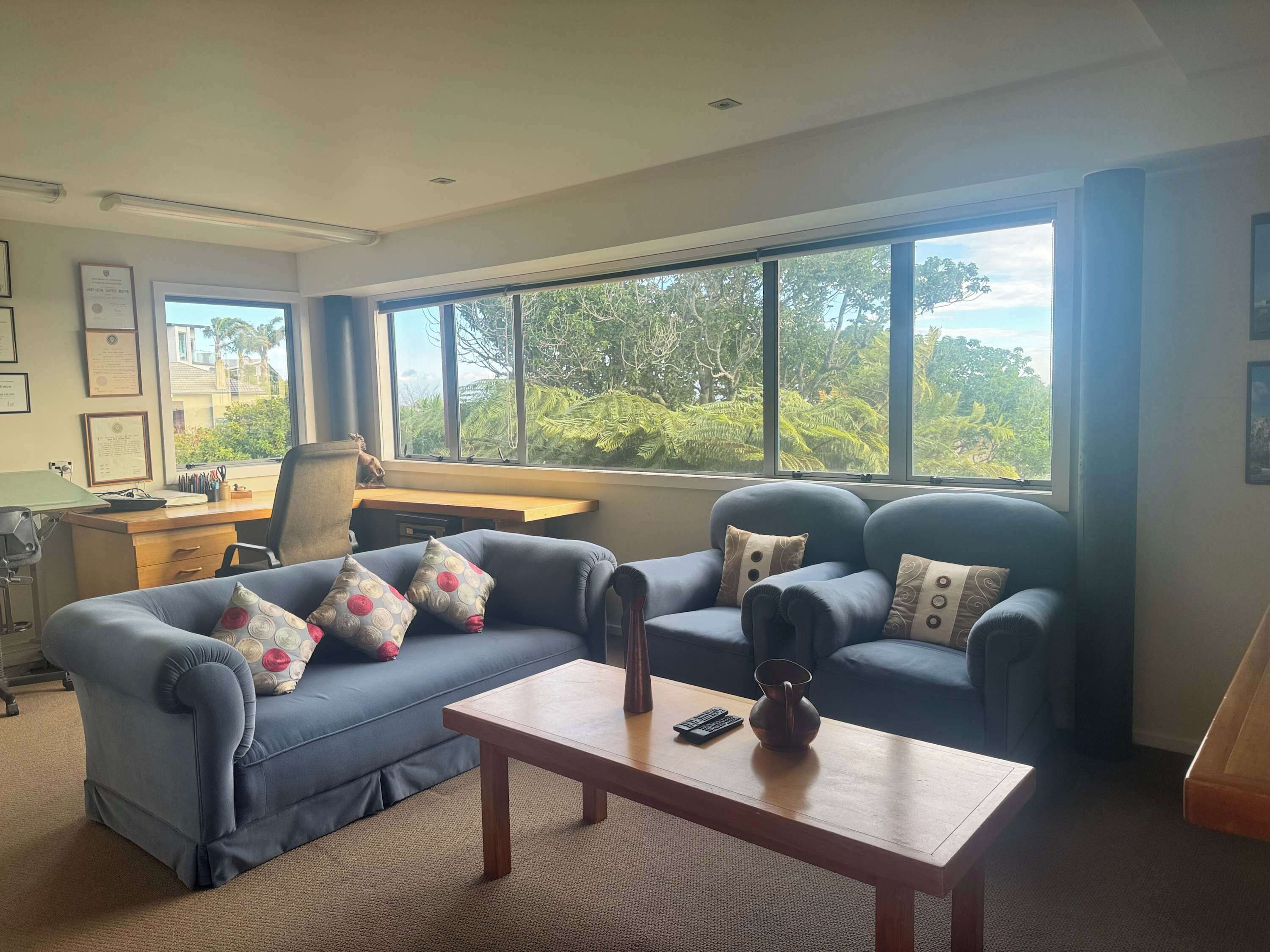 A living room with a sofa, a coffee table, and a desk with a computer. There are large windows with a view of trees and a building.