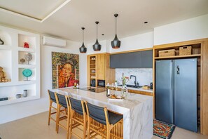 A modern kitchen with a marble island, wooden chairs, and a black refrigerator. - Villa Sukha Senja Bingin (Pecatu)