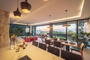 Un espace de salle à manger moderne, doté d’une grande table, de chaises et de luminaires suspendus.