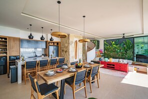 A modern kitchen with a dining area, wooden furniture, and a large window with plants. - Villa Sukha Senja Bingin (Pecatu)