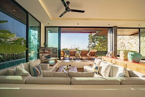 A modern living room with a large sofa, a ceiling fan, and a view of the outdoors. - Villa Sukha Senja Bingin (Pecatu)