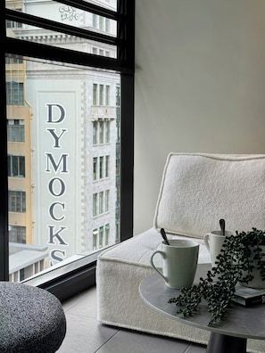 A modern room with a view of a building labeled 'DYMOCKS,' a white cushioned chair, a small round table with a mug and spoon, and a potted plant. - Sleek CBD Apartment | Steps to Everything Sydney (Sydney)