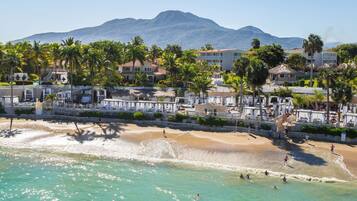 Hotel aan het strand met palmbomen, een zandstrand en een berg op de achtergrond.