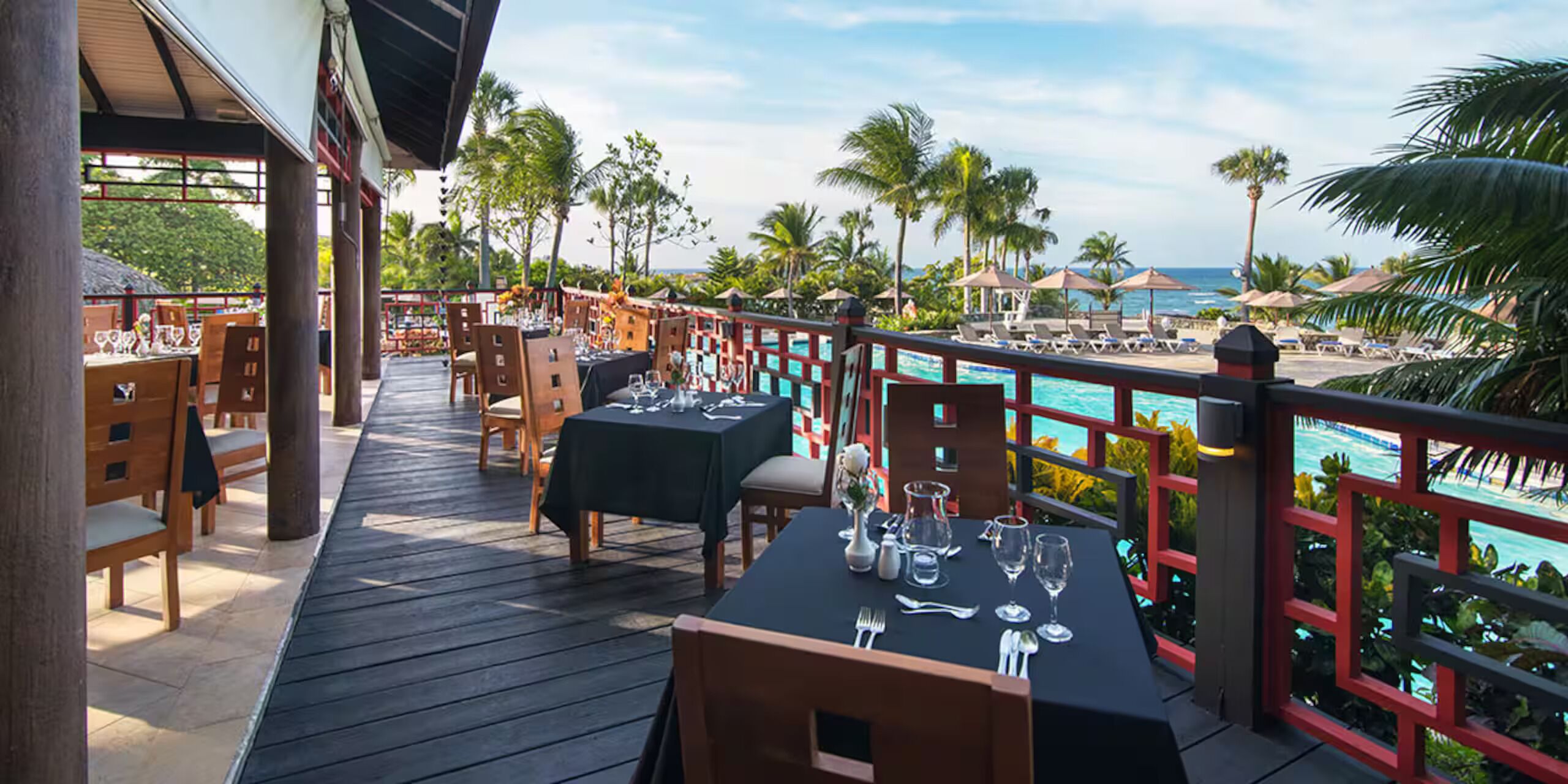 A deck with tables set for dining, overlooking a pool and palm trees.