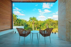 Een terras met twee rieten stoelen en een tafel, met uitzicht op een weelderig landschap met palmbomen en een helderblauwe lucht.