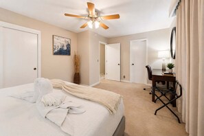 A hotel room with a bed, a desk, a chair, a ceiling fan, and a painting on the wall.