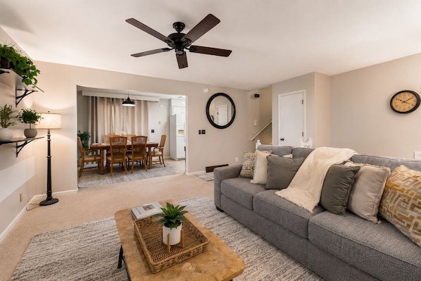A living room with a grey sofa, a coffee table, a dining area with a table and chairs, a ceiling fan, and a clock on the wall.