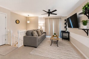 A living room with a sofa, a coffee table, a ceiling fan, a TV mounted on the wall, and a staircase with white railings.