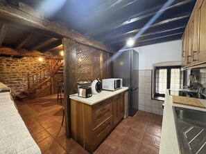 A rustic kitchen with a stone wall, wooden beams, a built-in stove, a refrigerator, and a window with curtains. - El Valle de la Reina House VV-4003-AS (Romillo)