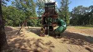 A playground with a wooden play structure, including a slide and climbing elements, set in a wooded area with a clear sky. - Cabin Near Thunder Ridge & Big Cedar_Private Hot Tub_Free Attraction Tickets_Bonus Loft (Ridgedale)