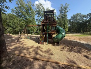 Ein Spielplatz mit einer Holzspielanlage, darunter eine Rutsche und Kletterelemente, inmitten einer bewaldeten Gegend mit blauem Himmel.