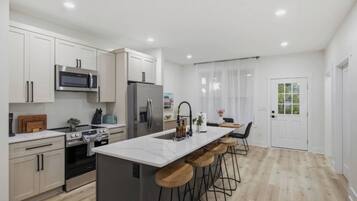A modern kitchen with white cabinetry, stainless steel appliances, a marble island with bar stools, and a dining area with a view of greenery.