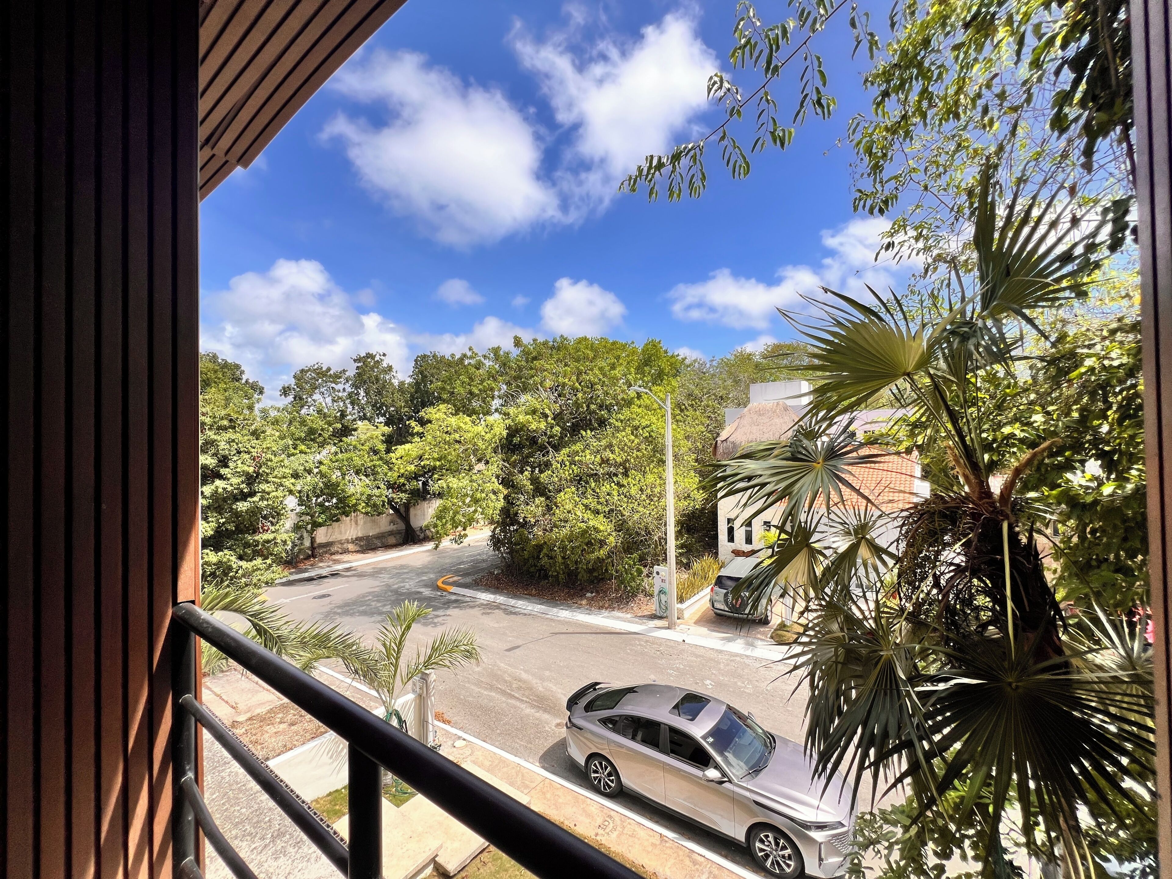 Une vue depuis un balcon donnant sur une rue où est garée une voiture, avec des arbres et un bâtiment dont le toit est recouvert de chaume.
