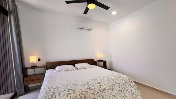 A bedroom with a bed, two bedside lamps, a ceiling fan, and an air conditioning unit.
