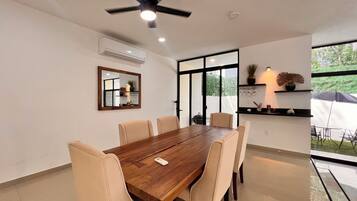 Uma sala de jantar com mesa de madeira e seis cadeiras, um ventilador de teto e uma janela grande com vista para o verde.