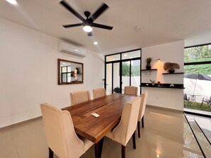 A dining area with a wooden table and six chairs, a ceiling fan, and a large window with a view of greenery. - Modern Elegant Villa | 3 Bedrooms • 4 BA | Puerto Morelos (Puerto Morelos)