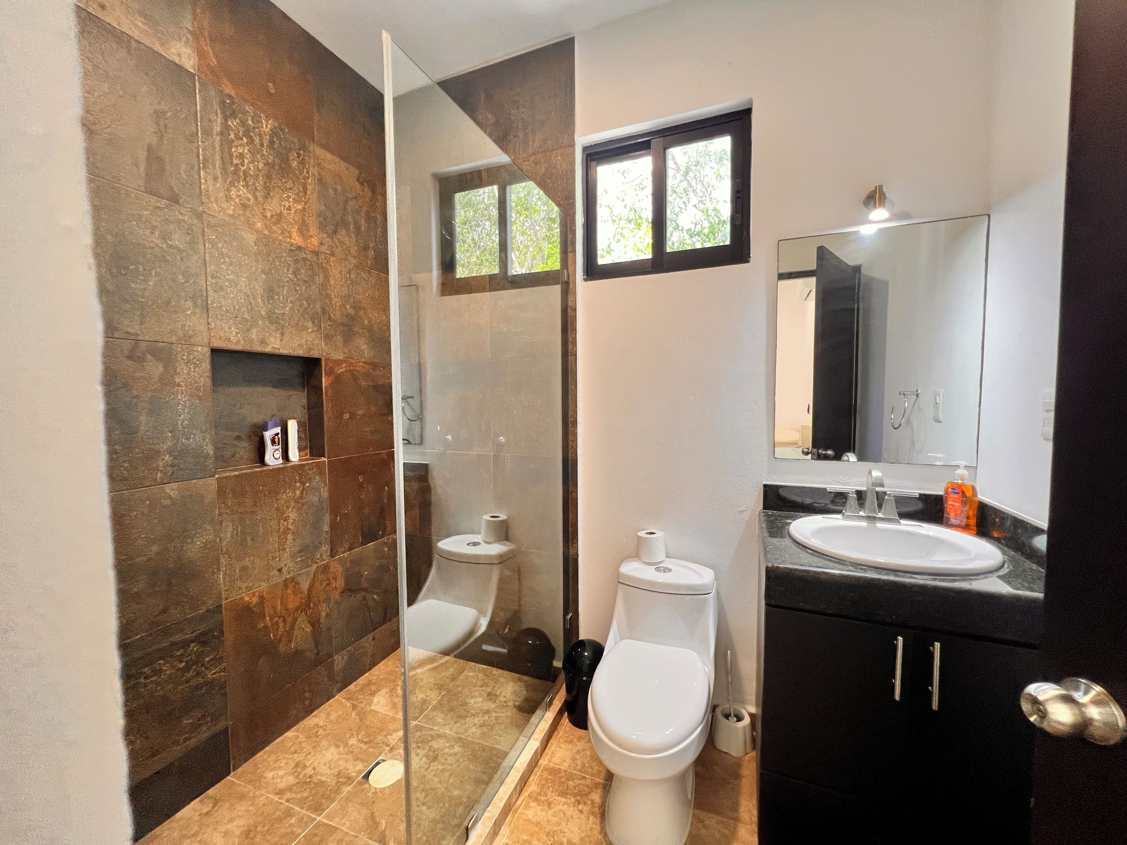 A bathroom with a glass shower enclosure, a toilet, a sink with a mirror, and a dark-colored cabinet.