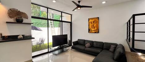 Una sala de estar moderna con un sofá seccional negro, un televisor de pantalla plana, un ventilador de techo y un ventanal con vistas a zonas verdes.