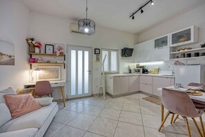 Uma cozinha moderna com armários brancos, uma área de jantar com mesa posta para dois e uma sala de estar com sofá e luminária.