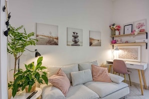 Ein Wohnzimmer mit einer Couch, einem Schreibtisch und einem Sessel. An den Wänden hängen Bilder und es steht eine Pflanze dort.