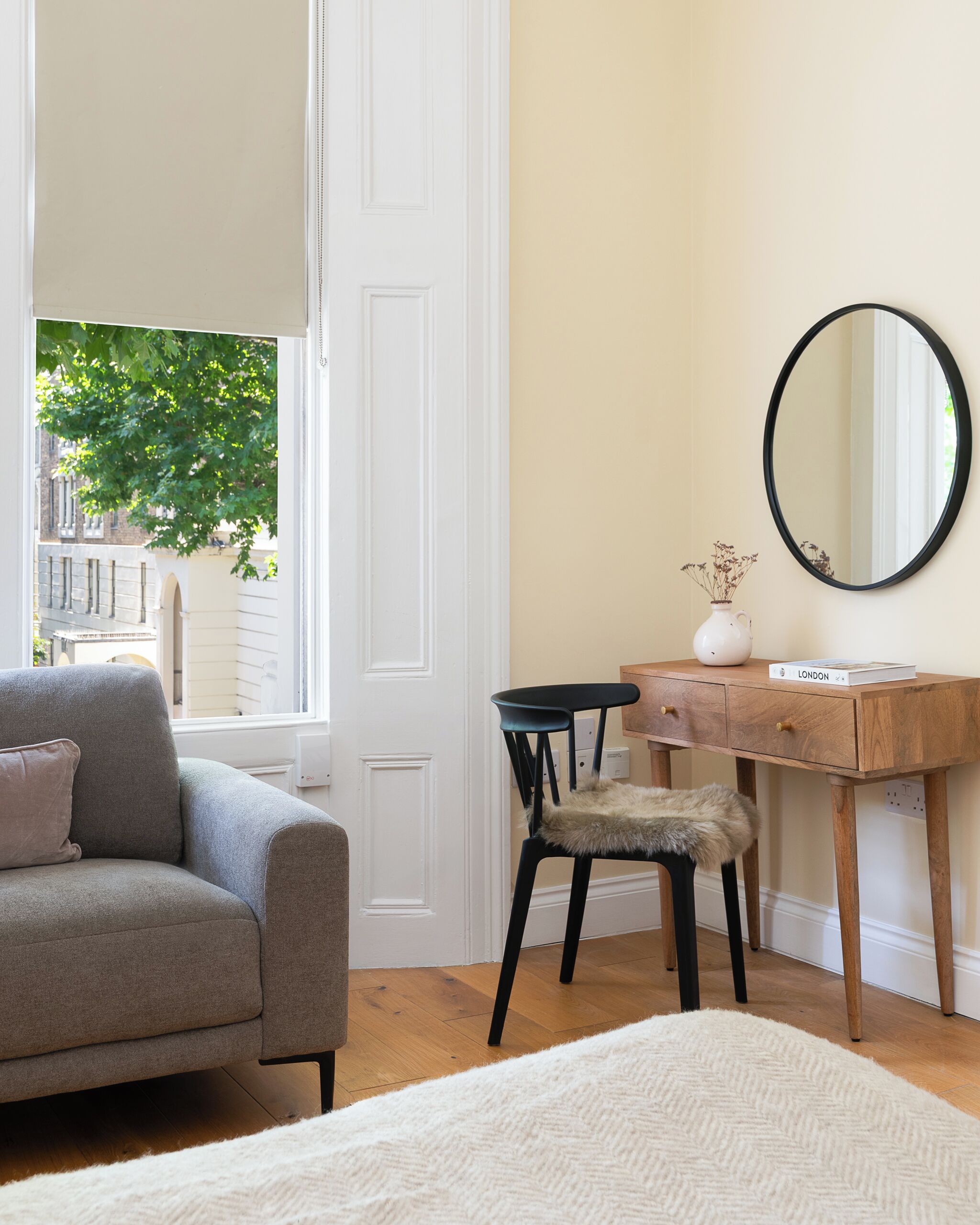 Ein Zimmer mit grauem Sofa, einem kleinen Holztisch, einem Stuhl, einem Spiegel und einem Fenster mit Blick auf Bäume und Gebäude.