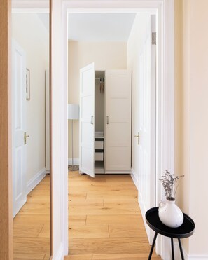 A hallway with a wooden floor leading to a room with a white door, a small table with a vase, and a closet with hanging items.