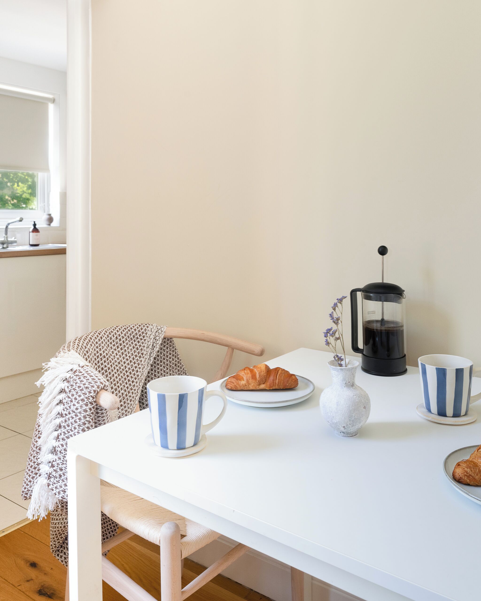 A white table with a coffee maker, two striped cups, a plate with croissants, and a vase with flowers.